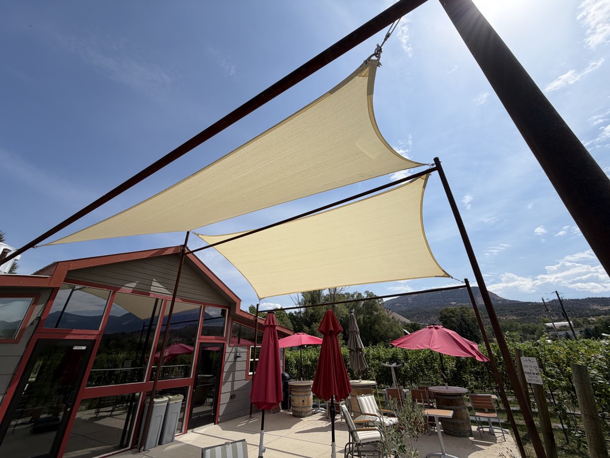 Two cream shade sails on steel posts over the Painted Vineyard tasting patio in Paonia, with red umbrellas and vines beyond